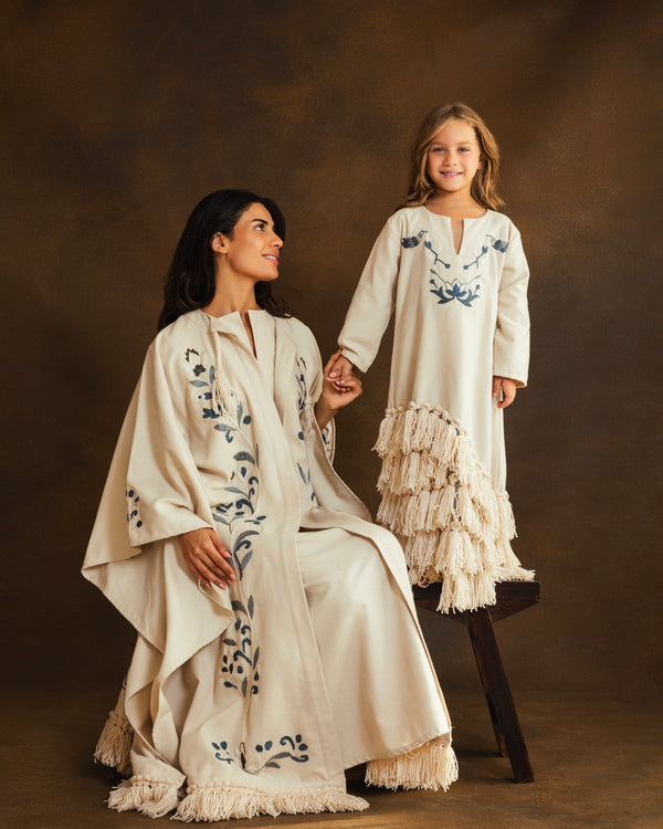 Mother-daughter wearing matching cream-colored garments with blue floral embroidery and fringe details, posed against a warm brown studio backdrop.