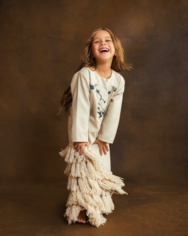 A young girl with long light brown hair is laughing joyfully while wearing a cream-colored long-sleeve dress with blue floral embroidery on the chest and a matching tiered fringe skirt, set against a warm brown textured studio backdrop.