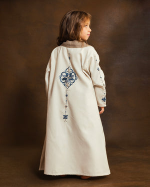 A young girl viewed from behind wearing a long cream-colored wool open kaftan with blue mosaic hand-embroidery on the back and sleeves, set against a dark brown studio backdrop.