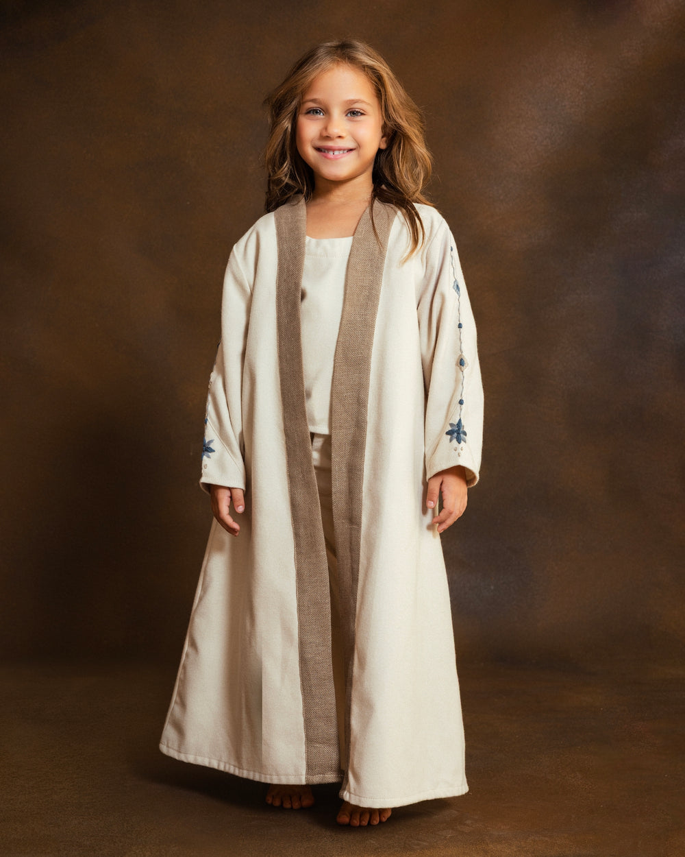 A young girl stands against a brown studio backdrop, wearing a long cream open kkaftan with a textured taupe-brown front panel and small blue hand embroidered mosaic details on the sleeves.