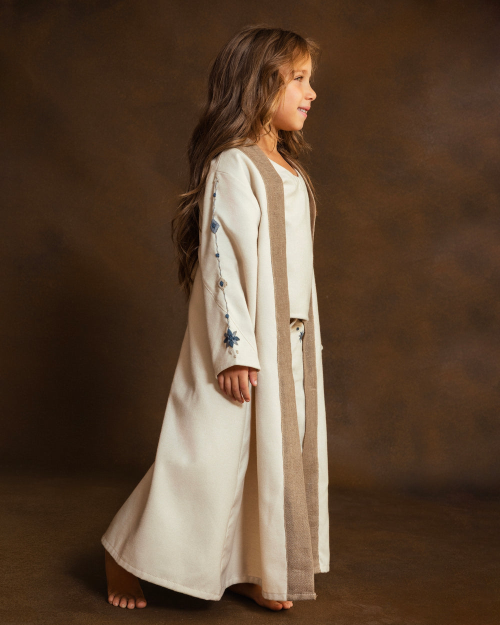 A young girl stands against a brown studio backdrop, wearing a long cream open kkaftan with a textured taupe-brown front panel and small blue hand embroidered mosaic details on the sleeves.