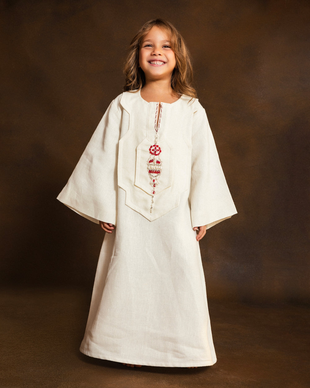 A young girl smiling while wearing a cream-colored linen kaftan with red embroidered details, set against a dark brown studio backdrop.