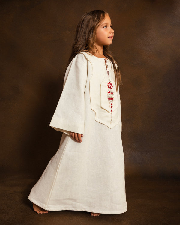A young girl with long brown hair stands in profile wearing a white linen kaftan with wide sleeves and red hand-embroidered details on the chest, posed against a dark brown studio backdrop.