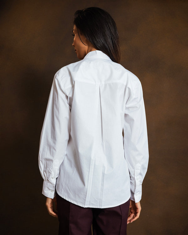 A woman shown from the back wearing a white long-sleeve button-up shirt and dark burgundy pants, standing against a dark brown studio background.