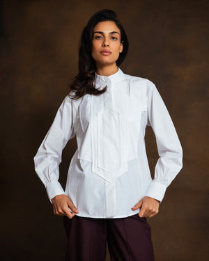 A woman modeling a white long-sleeve blouse with a high mock neck, gathered cuffs, and a large central pleated mosaic panel detail, paired with dark purple trousers, against a dark brown studio background.
