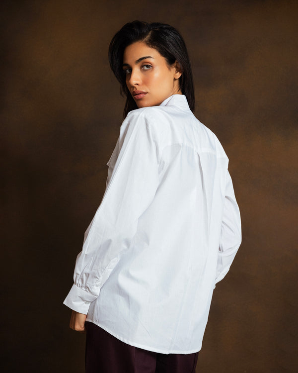 A woman wearing an oversized white long-sleeve button-up shirt, photographed from the back against a dark brown studio backdrop.