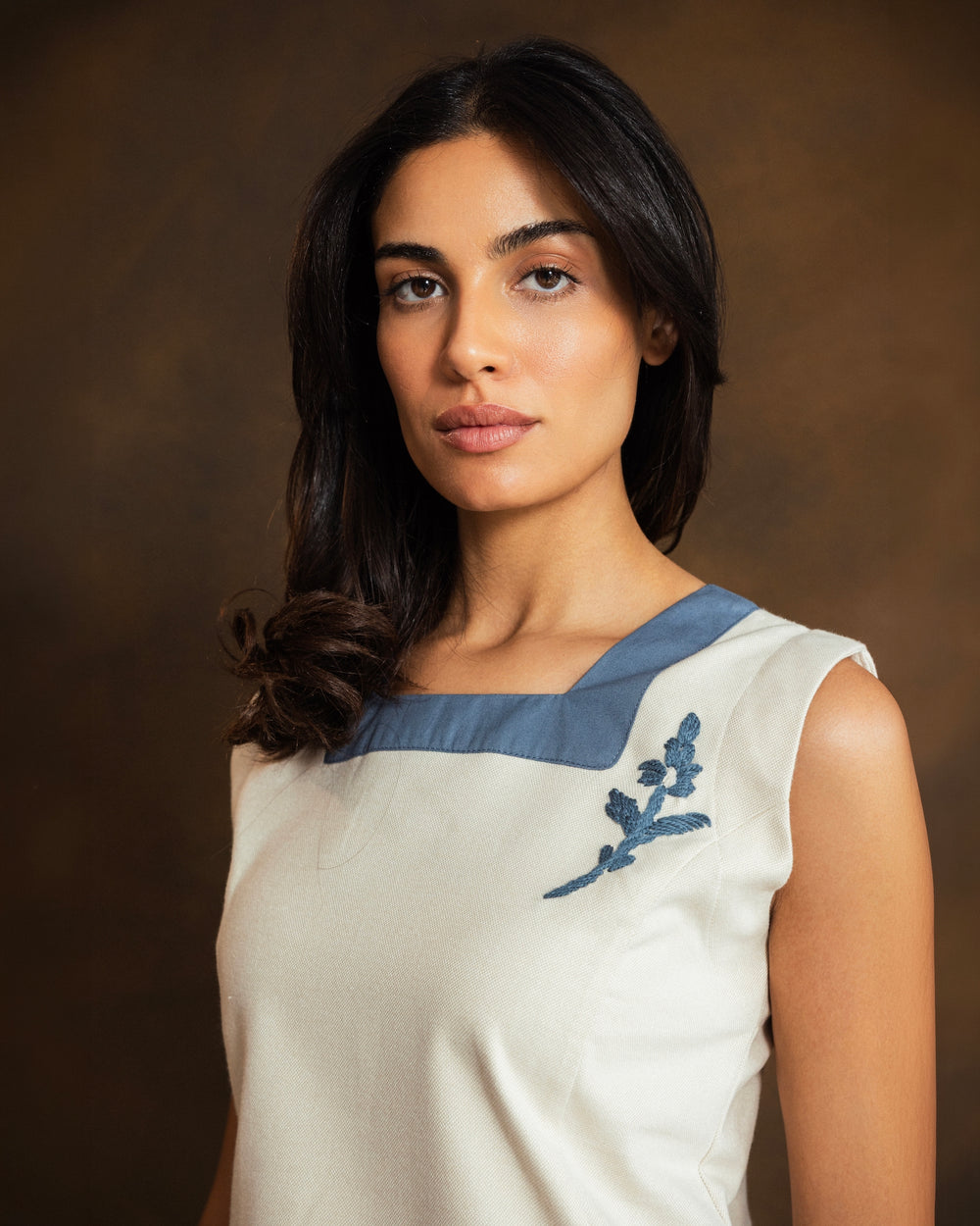 A woman modeling a sleeveless cream-colored top with a contrasting blue collar and a blue embroidered floral motif on the chest, paired with matching cream trousers, set against a dark brown textured studio backdrop.