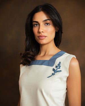 A woman with dark hair wearing a sleeveless cream-colored top with a muted blue collar and a blue floral embroidery on the chest, set against a dark brown studio background.