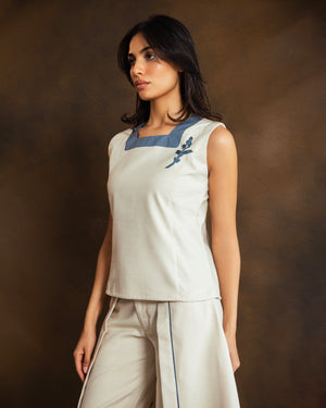 A woman modeling a sleeveless cream-colored top with a contrasting blue collar and a blue embroidered floral motif on the chest, paired with matching cream trousers, set against a dark brown textured studio backdrop.