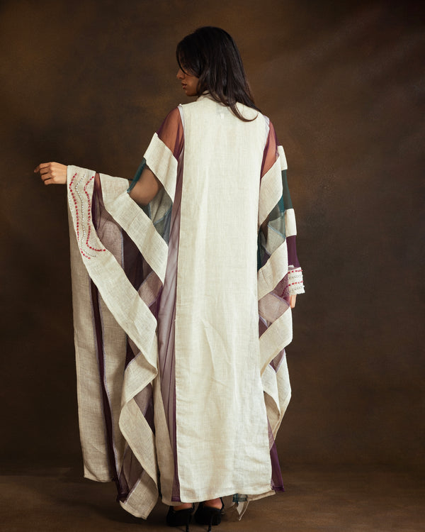 A woman shown from behind wearing a long, loose white linen caftan with wide sleeves featuring purple, green, and beige striped patterns, posed against a dark brown studio backdrop.