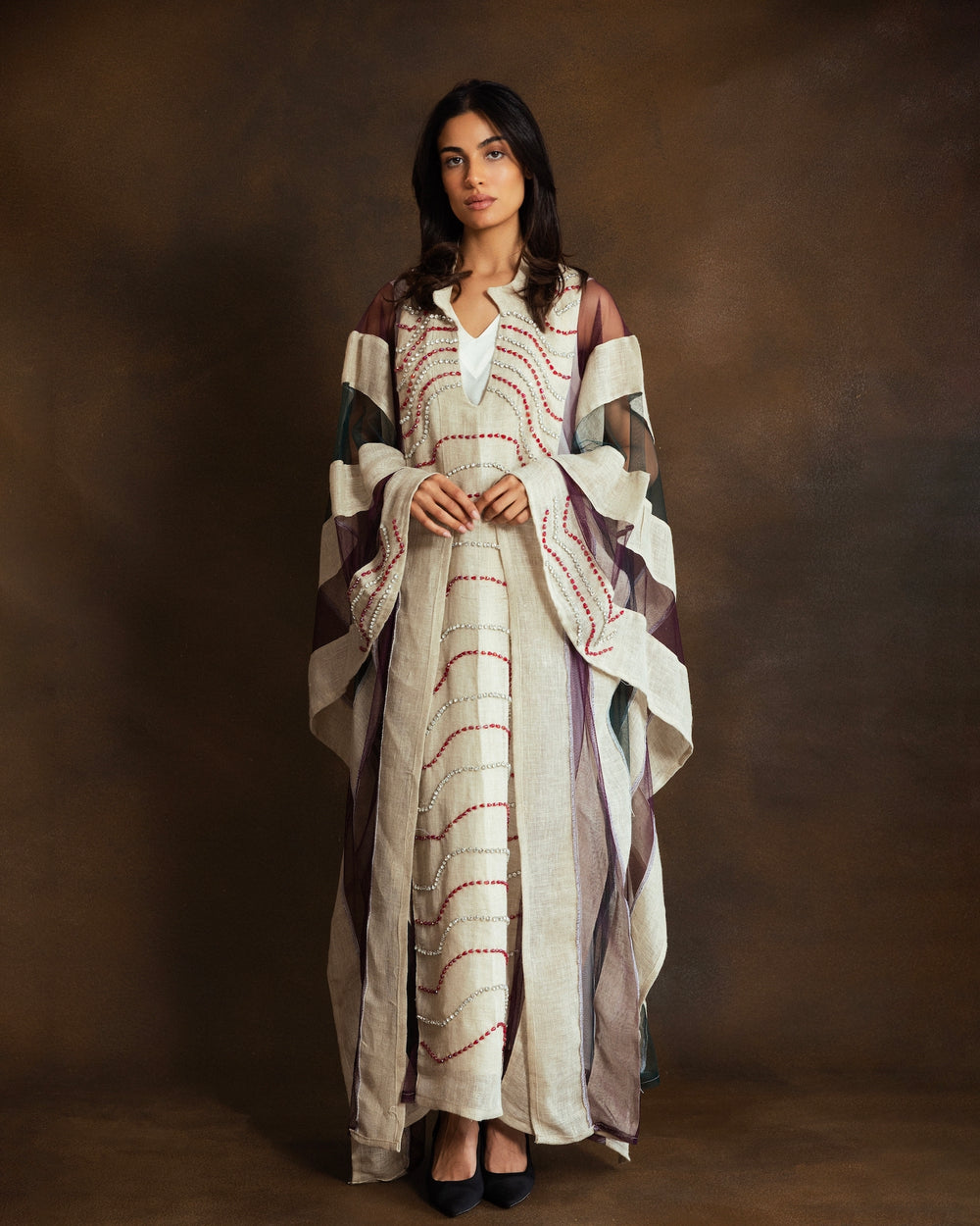 A woman stands against a warm brown studio backdrop wearing a long, loose stripes kaftan with wide draped sleeves and red embroidered curved lines. The dress has a V-neck and a sheer overlay on the sleeves.