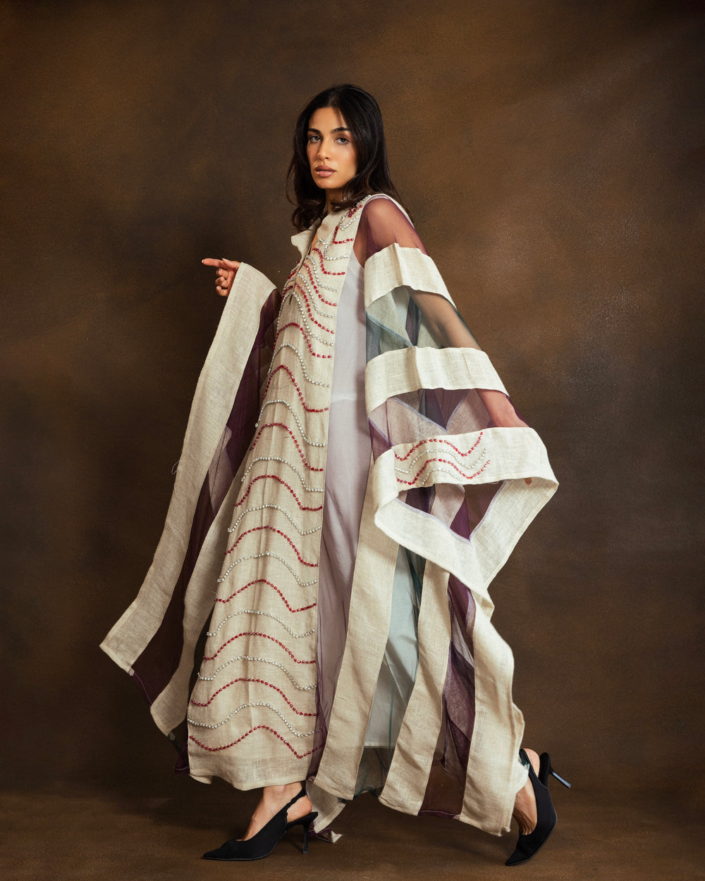 A woman stands against a warm brown studio backdrop wearing a long, loose stripes kaftan with wide draped sleeves and red embroidered curved lines. The dress has a V-neck and a sheer overlay on the sleeves.