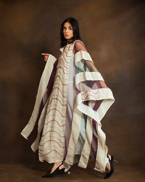 A model wearing a long, flowing cream-colored dress with red wavy embroidered lines and wide draped sheer sleeves, posed against a textured brown studio backdrop.