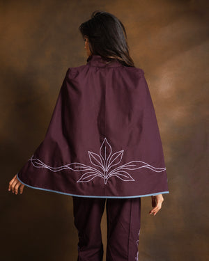 A woman shown from behind wearing a deep burgundy cape with dusty pink embroidered leaf details and a light blue trim, paired with matching pants, against a textured brown studio backdrop.