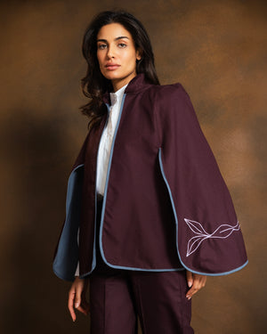 A woman models a deep plum purple cape-style jacket with light blue piping and white embroidered leaf details on the sleeve, paired with matching trousers and a white shirt, against a warm brown studio background.