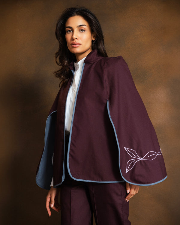 A woman models a deep plum purple cape-style jacket with light blue piping and white embroidered leaf details on the sleeve, paired with matching trousers and a white shirt, against a warm brown studio background.