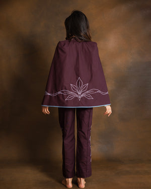 A woman viewed from behind wearing a dark plum purple poncho-style cape with dusty pink embroidered leaf details and matching pants, standing against a textured brown studio backdrop.