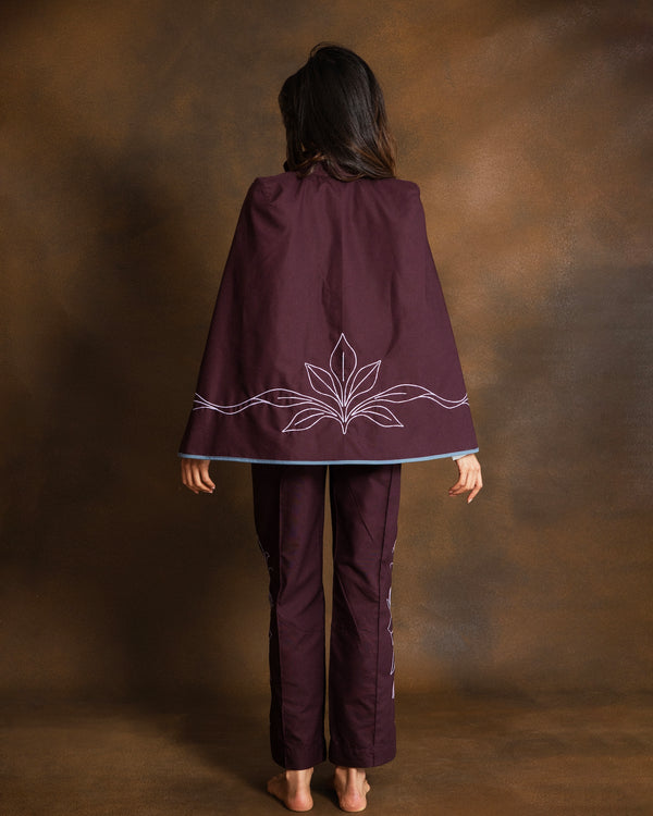 A woman viewed from behind wearing a dark plum purple poncho-style cape with dusty pink embroidered leaf details and matching pants, standing against a textured brown studio backdrop.