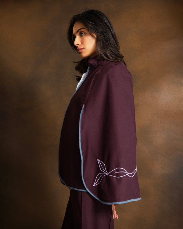 A woman wearing a deep plum-purple cape-style jacket with light blue trim and dusty pink embroidered leaf details on the sleeve, posed against a warm brown studio backdrop.
