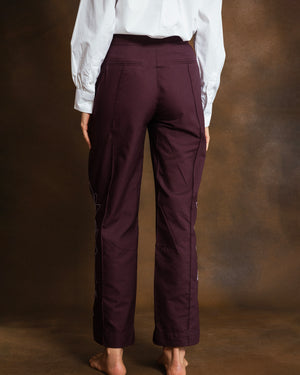 A person shown from the back wearing dark plum tailored pants and a white blouse, standing against a warm brown studio backdrop.