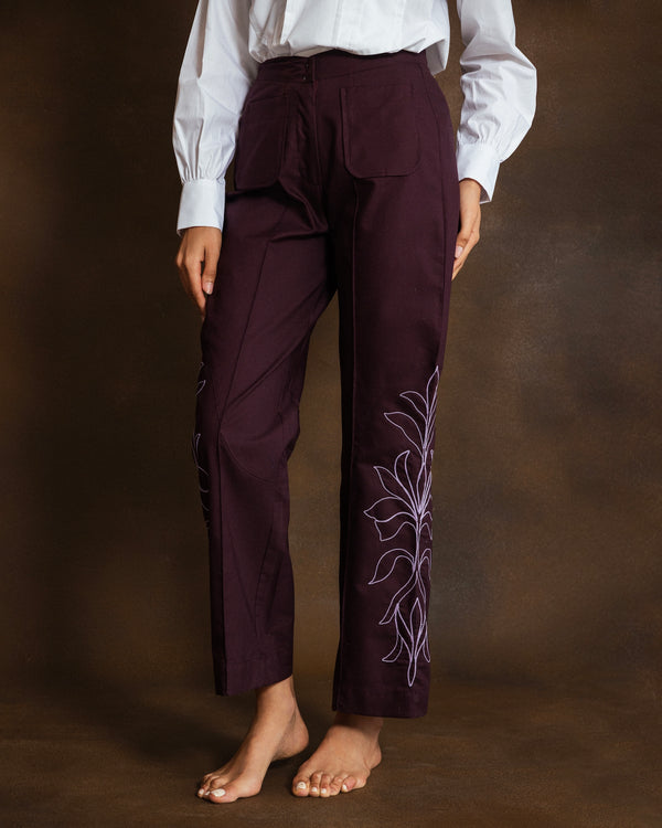 A person wearing dark plum pants with dusty pink floral embroidery on the lower leg, paired with a white long-sleeve blouse, standing against a dark brown studio background.