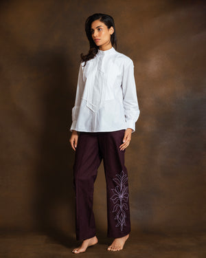 A woman stands against a textured brown studio backdrop, wearing a white long-sleeve blouse with a high gathered neckline and dark plum wide-leg pants featuring light purple floral embroidery on one leg.