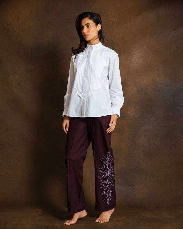 A woman stands against a textured brown studio backdrop, wearing a white long-sleeve blouse with a high gathered neckline and dark plum wide-leg pants featuring light purple floral embroidery on one leg.