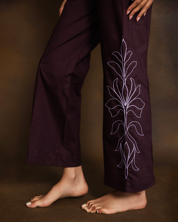 A close-up of a person wearing dark plum wide-leg pants with dusty pink floral embroidery down the outer side of one leg, shown against a warm brown studio background.