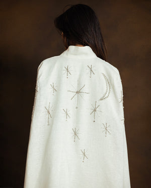 A person shown from behind wearing a white high-neck cape kaftan with embroidered celestial motifs, including stars and a crescent moon, against a dark brown background.