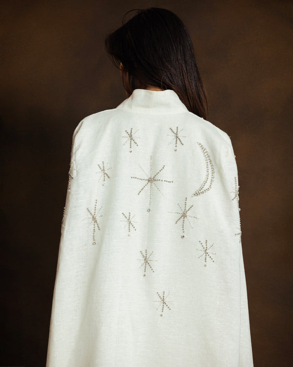 A person shown from behind wearing a white high-neck cape kaftan with embroidered celestial motifs, including stars and a crescent moon, against a dark brown background.
