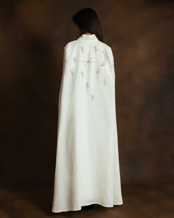 A person viewed from behind wearing a long white cape style kaftan with embroidered celestial motifs, including a crescent moon and stars, against a dark brown studio backdrop.