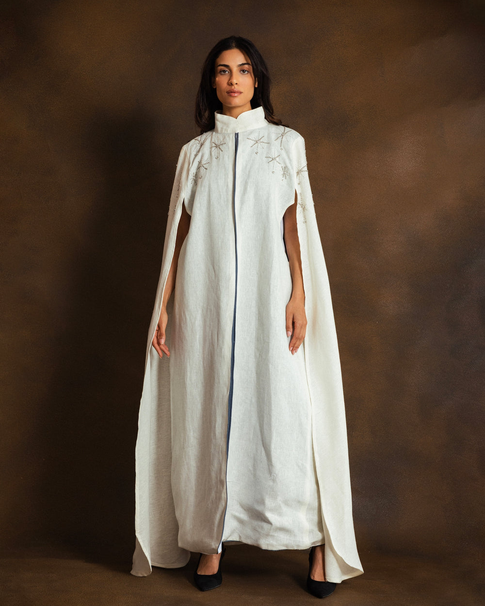 A woman modeling a long white cape-style dress with a high mock neck, side slits, and delicate silver embroidery on the chest, set against a dark brown textured studio backdrop.