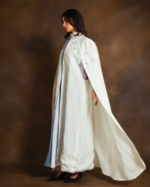 A woman models a long white linen open kaftan with delicate hand-embroidery, . She is posed in profile against a textured brown studio backdrop.