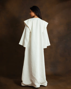 A woman wearing a long, flowing white kaftan with wide layered sleeves, shown from the back against a dark brown studio backdrop.