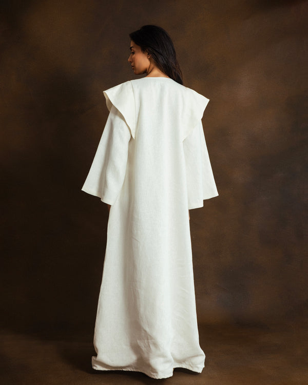 A woman wearing a long, flowing white kaftan with wide layered sleeves, shown from the back against a dark brown studio backdrop.