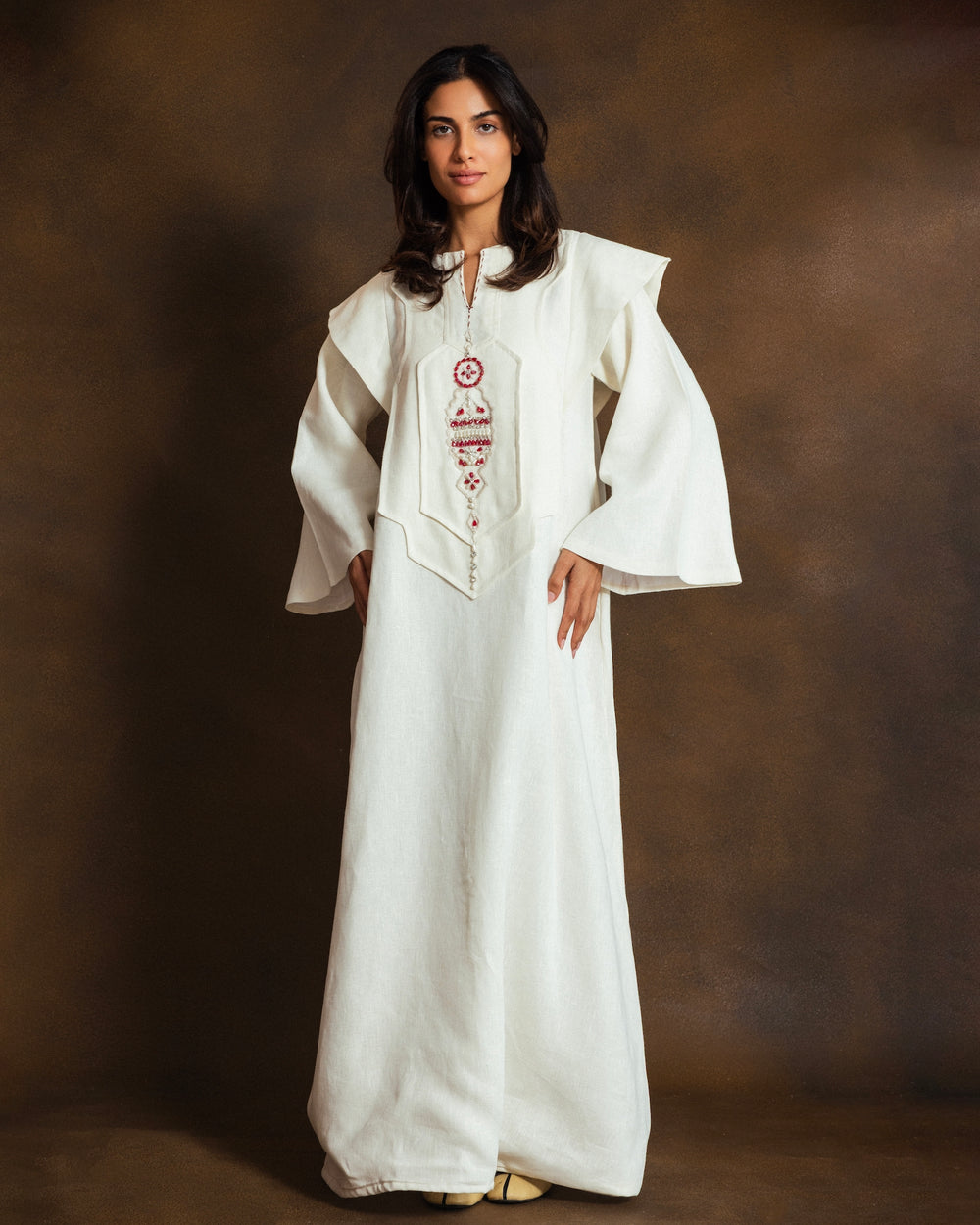 A woman modeling a long white traditional dress with wide flared sleeves and red embroidered detailing on the front, set against a dark brown studio backdrop.