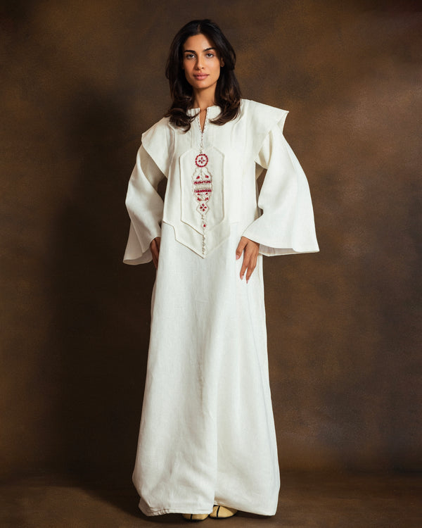 A woman modeling a long white traditional dress with wide flared sleeves and red embroidered detailing on the front, set against a dark brown studio backdrop.