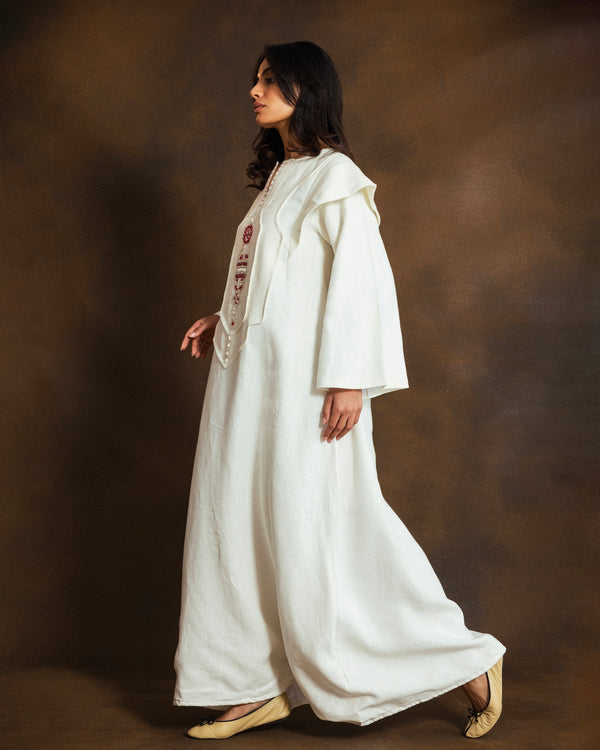 A woman wearing a long white linen Kaftan with red floral embroidery on the chest, standing in profile against a textured brown studio backdrop.