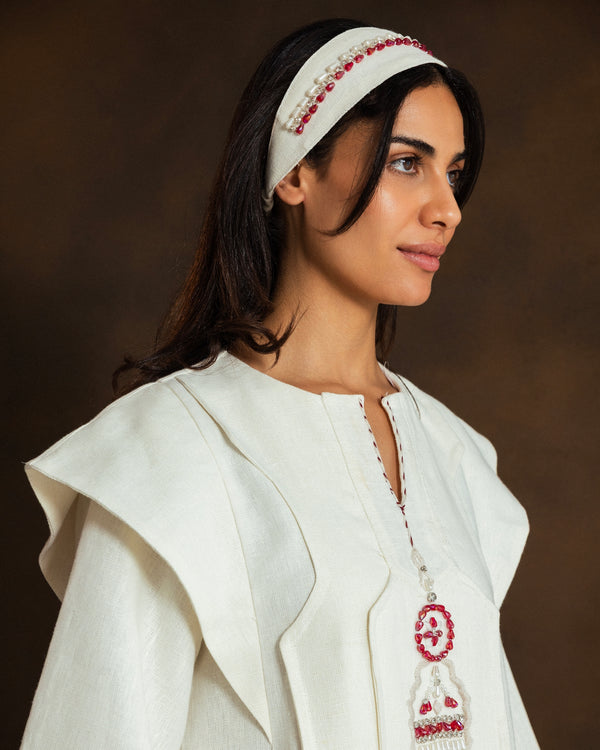 A woman wearing a cream-colored kaftan with red and white embroidered details, a matching cream headband with red beadwork, set against a dark brown studio background.