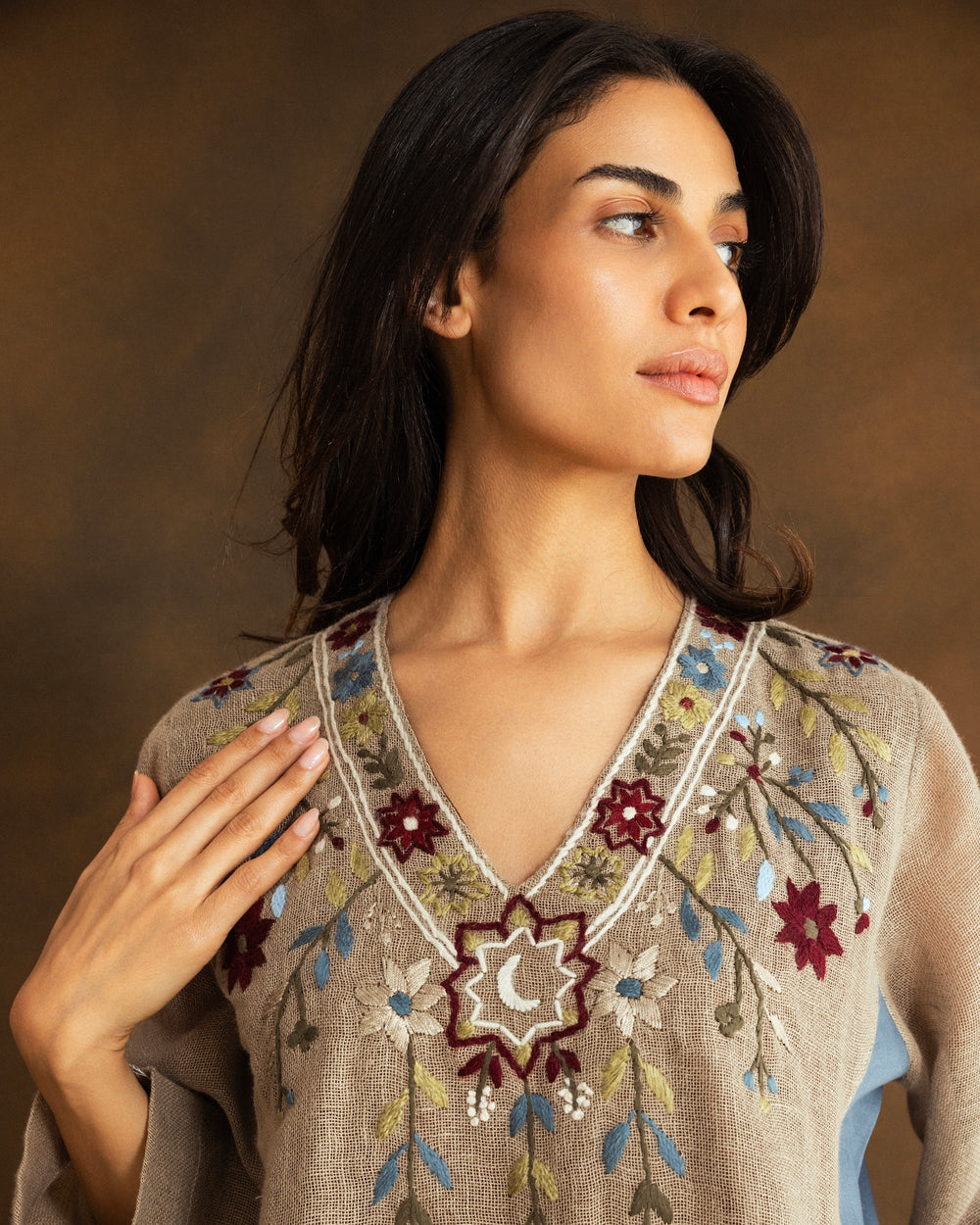 Woman wearing a beige raffia linen kaftan with hand-embroidered floral motifs, a crescent moon inspired by Ramadan nights, and contrasting dusty blue side panels.