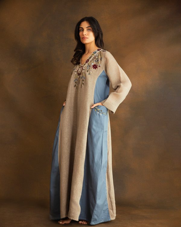 Woman wearing a beige raffia linen kaftan with hand-embroidered floral motifs, a crescent moon inspired by Ramadan nights, and contrasting dusty blue side panels.
