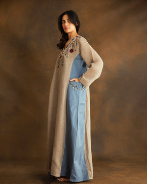 Side view of a woman wearing a beige raffia linen kaftan with dusty blue gabardine side panels, embroidered pockets and a flowing silhouette.