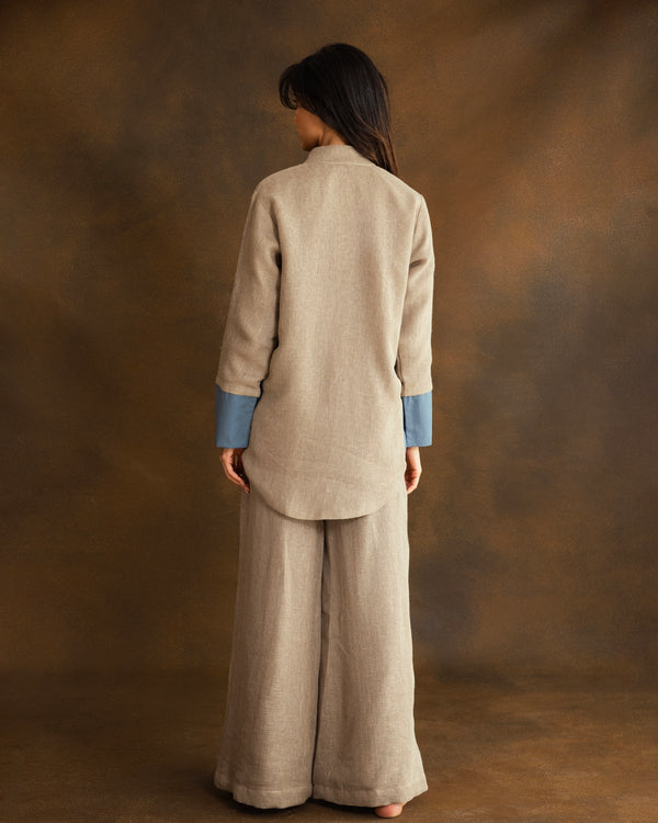 A model shown from the back wearing a beige linen two-piece set consisting of a long-sleeve top with contrasting blue cuffs and matching wide-leg trousers, set against a textured brown studio backdrop.