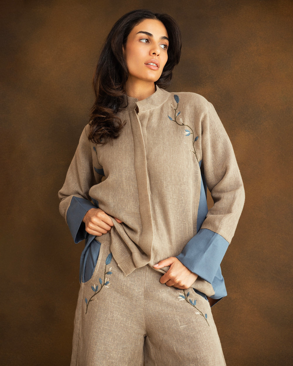 A woman modeling a beige linen two-piece set consisting of a long-sleeve button-front tunic with contrasting blue cuffs and matching wide-leg trousers, posed against a textured brown studio backdrop.