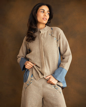 A woman modeling a beige linen two-piece set consisting of a shirt with blue contrast cuffs, side panels and embroidery, and matching trousers, against a textured brown studio background.