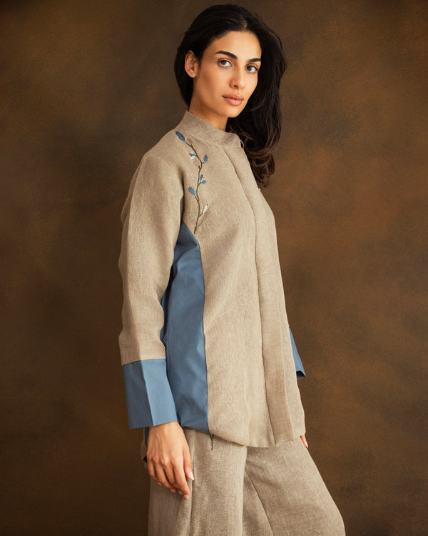A woman modeling a beige linen shirt with blue color-blocked panels on the sleeves and a small blue floral hand-embroidery on the chest, paired with matching beige trousers, set against a warm brown studio background.