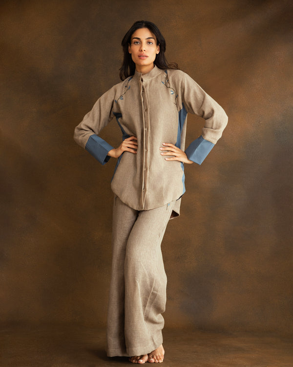A woman modeling a beige linen two-piece set with hand-embroidered floral detailing consisting of a long-sleeve button-front shirt with contrasting blue cuffs and matching wide-leg trousers, posed against a textured brown studio backdrop.