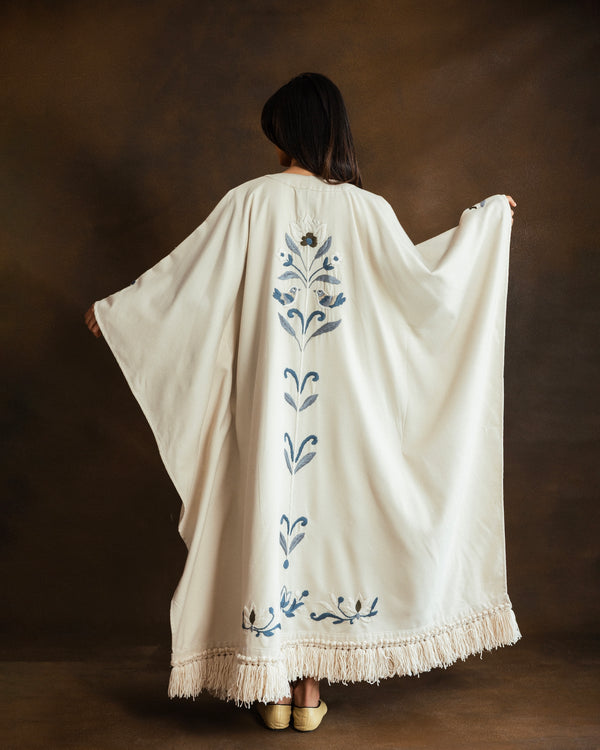 Back view of the YMZ Giardino Celeste Kaftan in cream wool blend, featuring delicate blue floral hand embroidery along the back and hem, photographed against a dark brown studio backdrop.