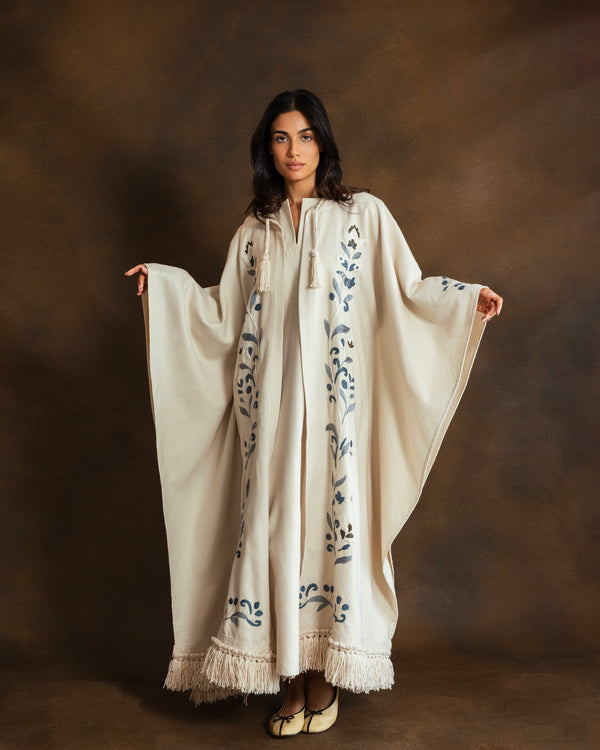 A woman modeling a cream-colored, wide-sleeved caftan or poncho with blue floral embroidery and a fringed hem, set against a dark brown studio backdrop.