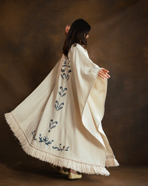 A woman wearing a flowing cream-colored kaftan with blue floral handembroidery and fringe trim, captured from behind as she twirls against a dark brown studio backdrop.